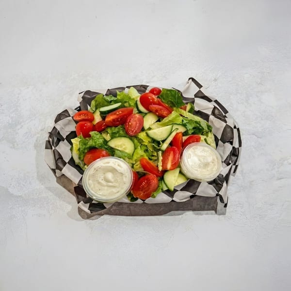 Fresh garden salad with lettuce, tomatoes, cucumbers, and two cups of dressing in a checkered paper-lined basket.