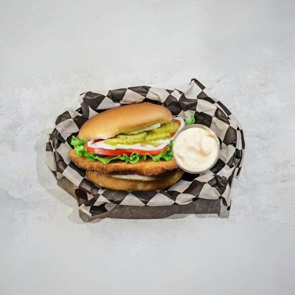 Crispy chicken sandwich with lettuce, tomato, onion, pickles, and a side of sauce in checkered paper.