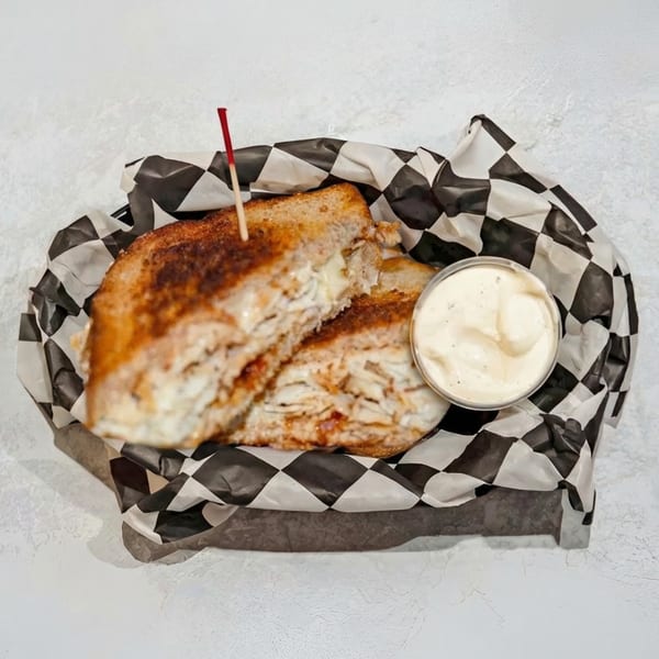Grilled sandwich with a side of dipping sauce, served in a basket with checkered paper.