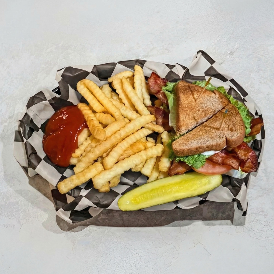 A club sandwich with fries, ketchup, and a pickle spear in a checkered paper-lined basket.